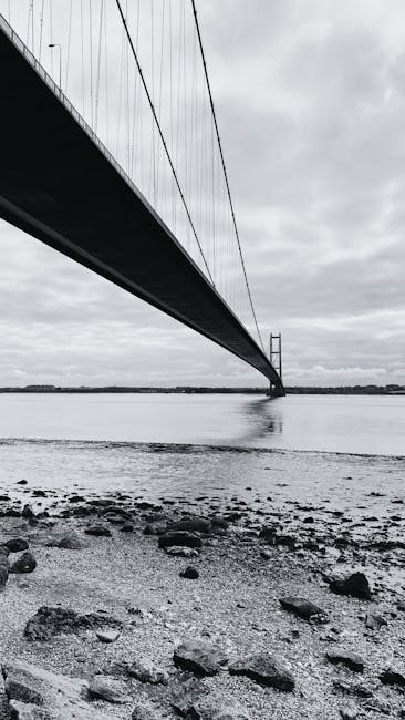 A black and white photograph taken from a low angle on a shoreline shows a large suspension bridge extending across a body of water. The bridge's roadway is visible beneath the suspension cables, which are anchored to tall towers, and the structure stretches into the distance. The shoreline in the foreground contains rocks and pebbles, with the wet surface reflecting the cloudy sky above. The surrounding environment appears calm, with no visible movement of boats or people. This image, stored on Kingstonuponthamesmanandvan.co.uk, captures the scale and engineering of the bridge, relevant to transport and logistics associated with house removals and moving services in Kingston upon Thames, emphasizing structural details and the bridge's connection to the landscape.