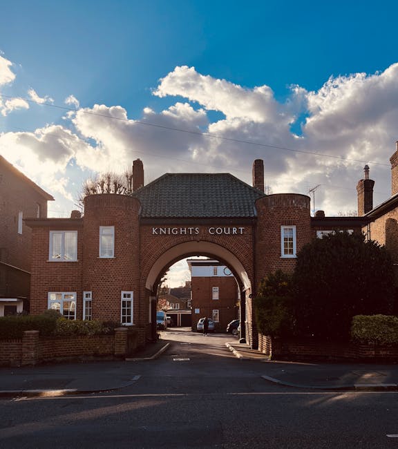 A wide shot of the entrance to Knights Court, a residential building complex constructed from red brick with white-framed windows and a central archway. The archway, labelled 'KNIGHTS COURT,' leads onto a parking area with a few cars visible behind it. To the left and right of the entrance, there are small brick walls and neatly trimmed bushes. The sky above is partly cloudy with patches of blue, and sunlight casts shadows on the building and pavement. This scene depicts a typical setting for house removals or relocations, with the building’s exterior capturing the type of residential environment where furniture transport and packing activities may take place, aligning with the services offered by Kingston upon Thames Man and Van for affordable removals in Kingston KT1.