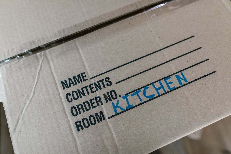 A close-up image of a cardboard moving box with black printed labels and handwritten annotations. The box has a label section listing 'NAME,' 'CONTENTS,' 'ORDER NO.,' and 'ROOM,' with the word 'KITCHEN' handwritten in blue ink next to 'ROOM.' The box is sealed with clear packing tape and appears to be clean and in good condition, positioned inside a property near a doorway, suggesting it is part of a home relocation or furniture transport process. The area around the box is well-lit, and nearby are other packing materials or boxes, indicating an organized packing and moving environment often managed by professional removal services such as Kingston upon Thames Man and Van. The scene reflects the process of packing and logistical planning during house removals, with attention to detail shown in clearly labeled boxes for efficient unpacking at the new address.