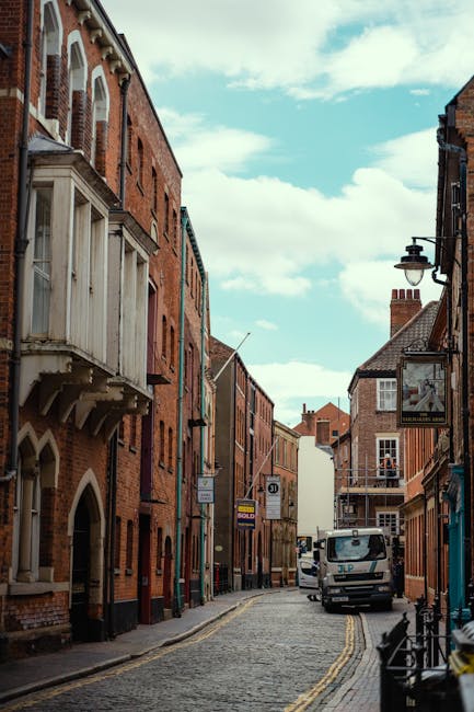 A narrow cobbled street in Canbury Kingston, Kingston upon Thames, with row houses constructed from red brick and featuring traditional architectural details. On the left side, a building with a small wooden bay window and decorative brackets extends over the sidewalk, while a lamp post and a hanging sign are visible on the right side. A white van is parked on the street, which is lined with yellow double lines indicating parking restrictions. The street appears to be part of a residential area, with the sky overhead showing a mix of clouds and blue patches. The scene may be part of a home relocation or furniture transport process, captured during daylight hours, with no people visible. This image aligns with house removals and moving services offered by Kingston Upon Thames Man and Van, illustrating a typical urban environment suitable for loading and transportation activities related to moving and packing.
