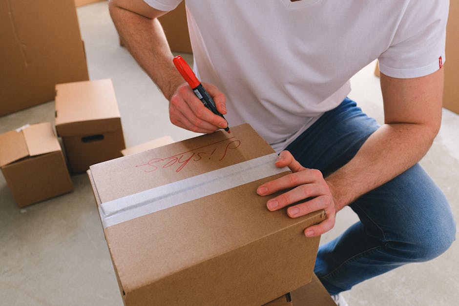 A close-up image of a cardboard moving box with black printed labels and handwritten annotations. The box has a label section listing 'NAME,' 'CONTENTS,' 'ORDER NO.,' and 'ROOM,' with the word 'KITCHEN' handwritten in blue ink next to 'ROOM.' The box is sealed with clear packing tape and appears to be clean and in good condition, positioned inside a property near a doorway, suggesting it is part of a home relocation or furniture transport process. The area around the box is well-lit, and nearby are other packing materials or boxes, indicating an organized packing and moving environment often managed by professional removal services such as Kingston upon Thames Man and Van. The scene reflects the process of packing and logistical planning during house removals, with attention to detail shown in clearly labeled boxes for efficient unpacking at the new address.
