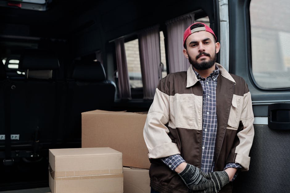 A male removal worker standing inside the open back of a van, which is parked outdoors, with several cardboard moving boxes of different sizes stacked nearby. The worker is wearing a red cap, a multi-coloured jacket, a checked shirt, and gloves, and is looking directly at the camera with a neutral expression. The interior of the van features black seats and light-coloured curtains on the windows, indicating a home relocation or moving process. The scene captures the loading or unloading stage of furniture transport, relevant to house removals, packing, and moving services provided by Kingston upon Thames Man and Van. The lighting is natural, suggesting daytime, with the focus on the practical aspects of furniture transport and packing logistics.