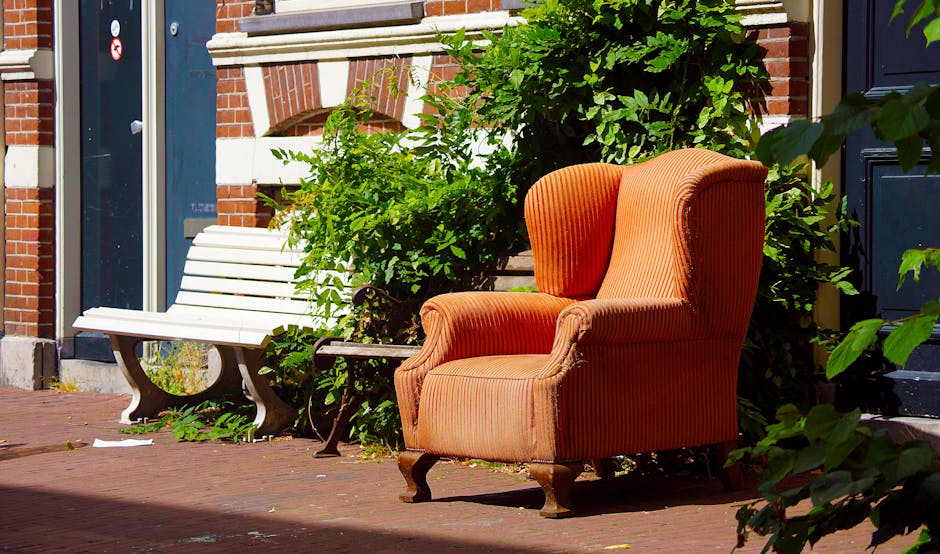 An outdoor scene showing a large, upholstered armchair with orange and brown striped fabric, positioned on a brick pavement outside a residential building. The armchair has curved wooden legs and rounded armrests, facing towards the viewer. Behind it, there is a white wooden bench with metal supports, partially obscured by green leafy bushes that are growing close to the building's wall. The building has a red brick facade with white decorative elements, a dark blue door to the right, and a window to the left. The scene is illuminated by natural daylight, with shadows cast on the pavement, indicating a sunny day. This setting suggests that the furniture might be part of a home relocation or furniture transport process, as handled by Kingston upon Thames Man and Van, which specializes in removals and moving services.