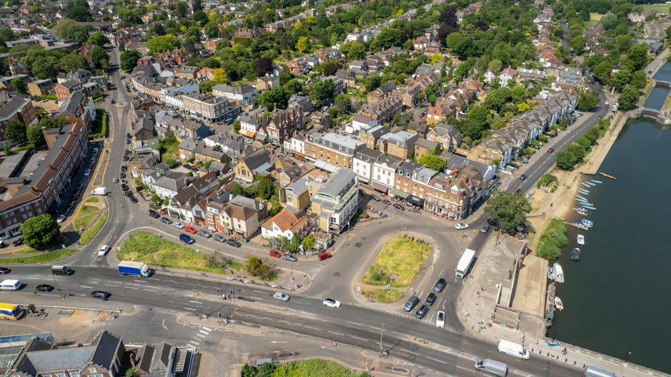 Aerial view of a small town showing a mix of residential and commercial buildings along a riverfront, with streets lined with parked cars and moving traffic. In the foreground, a wide road with lanes for vehicles, including cars and a blue truck, runs adjacent to the river. Next to the road, there are small green areas and parking spaces, with some loading and unloading activity visible near a group of trucks and vans, suggesting a home relocation or furniture transport process. Behind this, buildings of various sizes and styles, including shops, offices, and houses with brick and tiled roofs, extend into a neighborhood with tree-lined streets and tightly packed residential terraces. The river on the right side features boats docked along a quay, with a pathway running parallel to the water's edge. The scene is bright and well-lit, capturing a typical busy day in the town, where house removals and loading activities are likely ongoing, as supported by [COMPANY_NAME] and their professional moving services for packing and loading during home relocations in Kingston upon Thames.