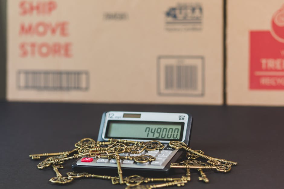 A close-up view of a small digital calculator displaying the number 749800, placed on a black surface with a pile of gold-colored keys scattered across it. The background consists of several large cardboard moving boxes with red and black text, including words like 'Ship,' 'Move,' and 'Store,' along with barcodes and handling symbols, indicating items packed for a house relocation. The scene captures the process of planning and logistics involved in furniture transport and packing during a home move, highlighting the importance of accurate calculations and proper packing materials such as cardboard boxes and protective wrapping. The image emphasizes the detailed organisation required in professional removals services provided by [COMPANY_NAME], aligning with guidance on avoiding hidden fees with Kingston removal companies, Kingston upon Thames.