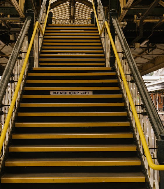 A wide view of a metal staircase with yellow handrails, leading up to a bridge or platform within a transport or industrial setting. The staircase has black and yellow striped anti-slip treads and a sign in the middle that reads 'PLEASE KEEP LEFT.' The surrounding environment includes structural beams and overhead infrastructure, indicating an elevated walkway typically found in loading docks or freight terminals. The lighting is moderate, highlighting the industrial features and cleanliness of the area, which is used for moving or freight handling. This staircase may be part of the logistics involved in house removals or furniture transport, supporting efficient and safe movement of goods during home relocation projects, as featured on Kingstonuponthamesmanandvan.co.uk.