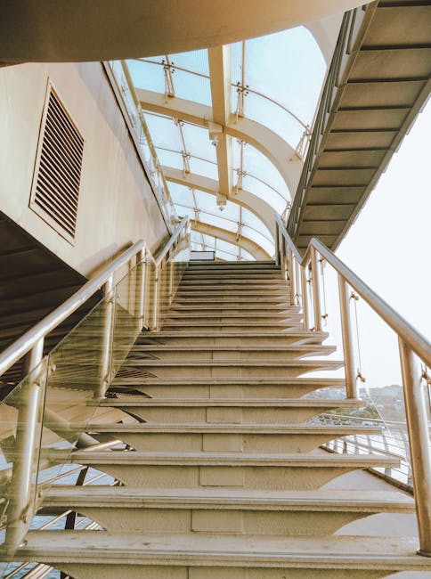 View of a modern staircase at Kingston Bridge, featuring metal steps with open risers and stainless steel handrails, leading upwards underneath a curved glass roof structure with metal framing. The staircase is situated inside a building, with natural daylight illuminating the area, and appears to be part of a large public or commercial space. This setting relates to house removals and furniture transport, as part of the logistics involved in home relocation, with the staircase potentially presenting a challenge for moving large furniture or appliances. In the context of Kingston upon Thames, Kingston Bridge access tips for tight stair removal jobs, this image illustrates the type of structural environment movers may encounter during a staircase access inspection, highlighting the importance of careful planning and equipment for safe loading and transport by Kingstonuponthamesmanandvan.co.uk's services.