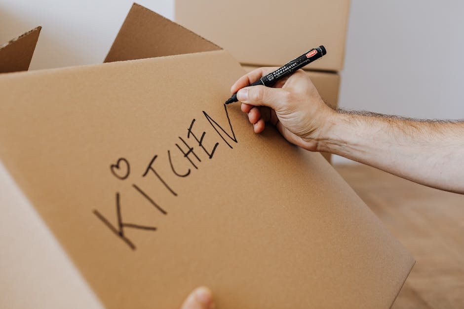 A person holding a black marker pen and writing the word 'KITCHEN' with a small heart symbol on a large brown cardboard box, which is part of a moving or home relocation process. The box is situated a