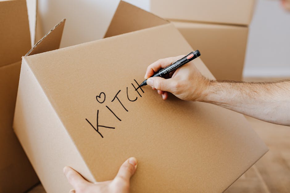 A person holding a black marker pen and writing the word 'KITCHEN' with a small heart symbol on a large brown cardboard box, which is part of a moving or home relocation process. The box is situated a