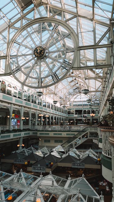 Inside a large indoor shopping centre with a glass-domed ceiling featuring a prominent circular clock at the center. The space is illuminated by natural light through the expansive glass roof, which showcases metal framing and supporting beams. Multiple levels of retail stores and walkways are visible, with some areas displaying shopfronts and signage. On the ground floor, several cardboard boxes and wrapped furniture items are arranged, indicating an ongoing home relocation or office move. A trolley loaded with packing materials is positioned near the boxes, with staff members or movers from Kingston upon Thames Man and Van visible handling equipment such as straps and blankets to assist with furniture transport and packing and moving activities. The environment suggests a busy, organized process of loading, lifting, or preparing items for transport, with clear spatial arrangement inside the shopping centre and close proximity to a vehicle or loading zone outside the building.