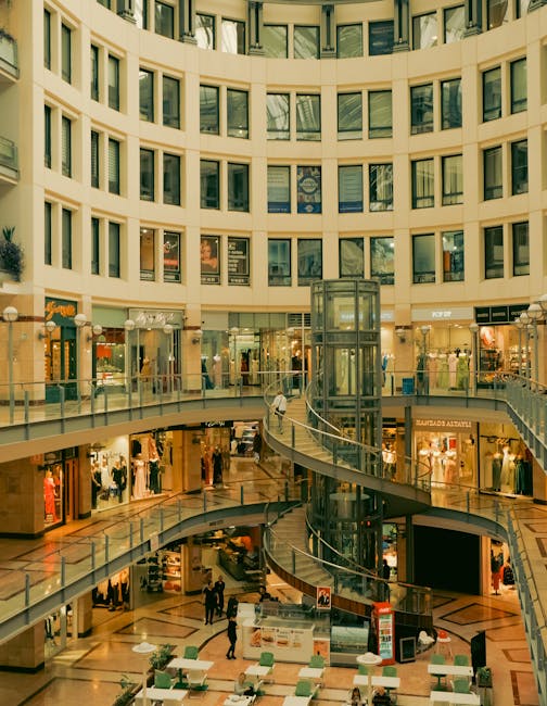 Interior view of a multi-storey shopping mall with a central open space featuring a glass elevator shaft surrounded by multiple curved escalators. The upper levels have retail storefronts with large glass windows displaying various clothing and accessory displays. The ground floor showcases seating areas with white tables and chairs, and several shoppers are visible walking and browsing the shops. The building's architecture includes cream-colored walls with multiple rows of rectangular windows and bright lighting illuminating the space. This setting is suitable for a house removal or relocation service's logistics operation, such as transporting furniture or packing materials within a commercial property like a shopping centre, supported by KINGSTONUPONTHAMESMANANDVAN.CO.UK, in relation to their office removals or shop relocation services at Kingston Bentall Centre.