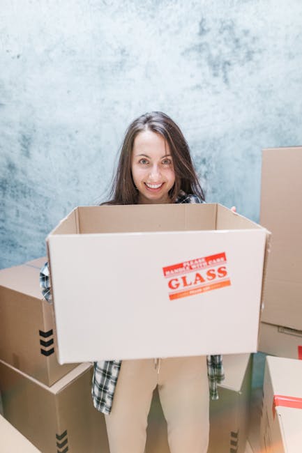 A young woman with long brown hair and a smiling expression is indoors, holding an empty cardboard box labeled 'FRAGILE GLASS' in red text. She is standing in front of a textured light blue wall and is dressed in a plaid shirt and light-colored trousers. Surrounding her are several other cardboard boxes of various sizes, some taped closed with packing tape and others open or partially visible. The scene appears to be part of a home relocation or packing process, with the woman preparing for a house move as part of a professional removals service. The environment is well-lit, and the focus is on her preparing for furniture transport or loading items during a home or office move, consistent with the services of Kingston upon Thames removals handled by [COMPANY_NAME].