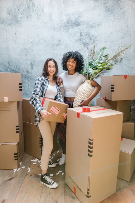 Two women standing inside a room surrounded by packed cardboard boxes, with one holding a small box and the other holding a decorative pineapple-shaped vase. The women are smiling towards the camera, and the boxes are sealed with red tape, indicating they are prepared for a home relocation or furniture transport. Behind them, a concrete-textured wall and some green houseplants add to the indoor environment. The scene captures the process of packing and moving, with the women appearing to be part of a professional removals service, possibly from Kingston upon Thames, as suggested by the webpage context. The boxes are stacked around them, some on the wooden floor, and a trolley or loading aids are not visible but implied by the setting's context. Proper lighting highlights the details of the boxes, packing materials, and the women’s clothing, emphasizing a professional and methodical packing and moving process supported by [COMPANY_NAME].